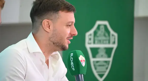 El entrenador del Elche CF, con camisa blanca, habla a un micrófono verde con el escudo del club.