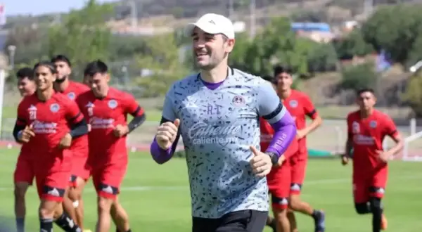 Entrenador sonriente lidera a jugadores en un partido pretemporada. Corren en un campo verde bajo el sol.