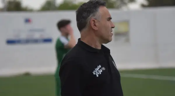 Entrenador de fútbol con polo negro, mirando hacia la derecha, con un jugador de verde al fondo.