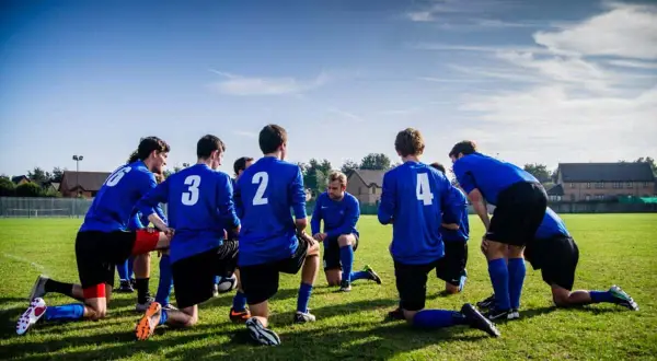 Equipo de fútbol con camisetas azules, arrodillados en el campo, preparándose para juegos de activación.