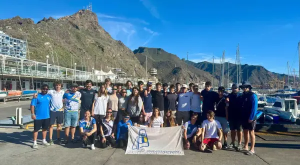 Grupo de jóvenes y adultos posan con una bandera del **CD Urda** en un puerto deportivo, con montañas y barcos de fondo.