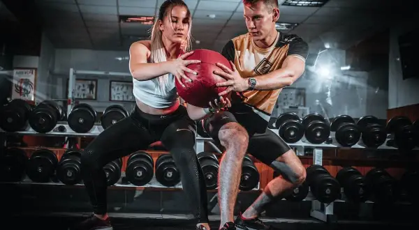 Pareja realizando ejercicios en pareja con pelota en un gimnasio, rodeados de pesas.