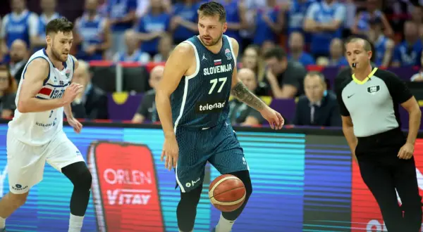 Jugador esloveno con el número 77 dribla el balón, mientras un árbitro observa atentamente. El jugador islandés, con el número 7, se prepara para defender. Se percibe una falta en ataque en el baloncesto.