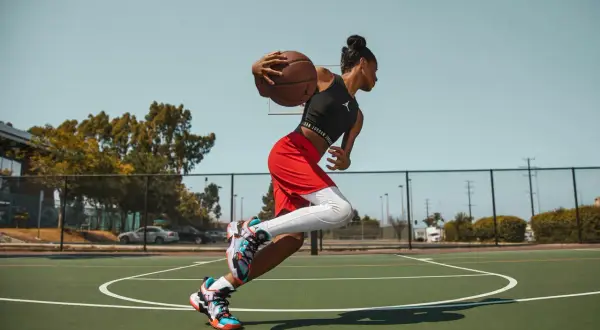 Jugadora de baloncesto en movimiento, con el balón en las manos, lista para anotar. Sus aleros de baloncesto brillan bajo el sol.