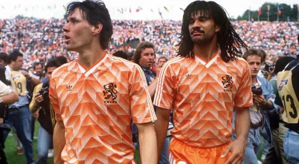 Dos futbolistas de equipos holandeses con camisetas naranjas icónicas, rodeados de aficionados y cámaras.