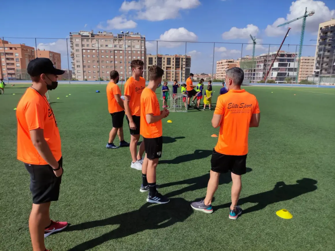 Entrenadores con camisetas naranjas dirigen a jóvenes deportistas en un campo de fútbol. Uno de ellos, con gorra negra, parece estar explicando algo, quizás sobre la equivalencia de técnico deportivo nivel 1.