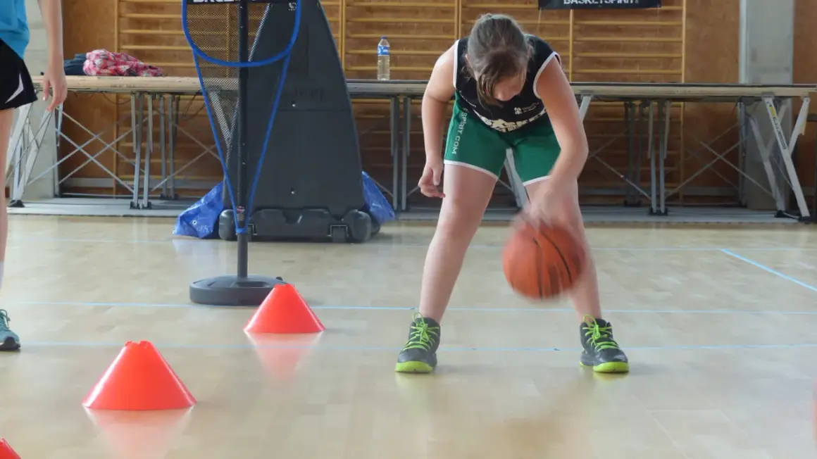 Niña concentrada botando el balón de baloncesto entre conos naranjas en un gimnasio.