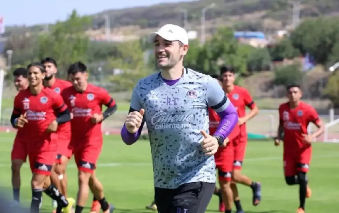 Entrenador sonriente lidera a jugadores en un partido pretemporada. Corren en un campo verde bajo el sol.