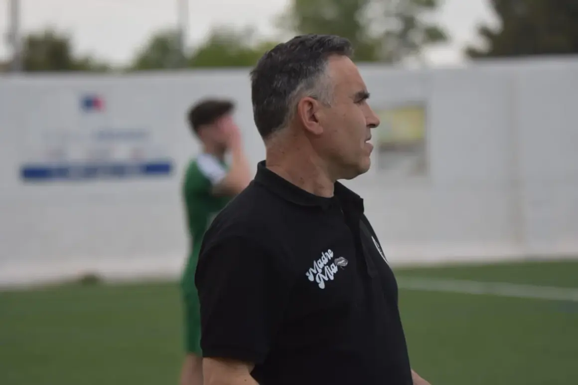 Entrenador de fútbol con polo negro, mirando hacia la derecha, con un jugador de verde al fondo.