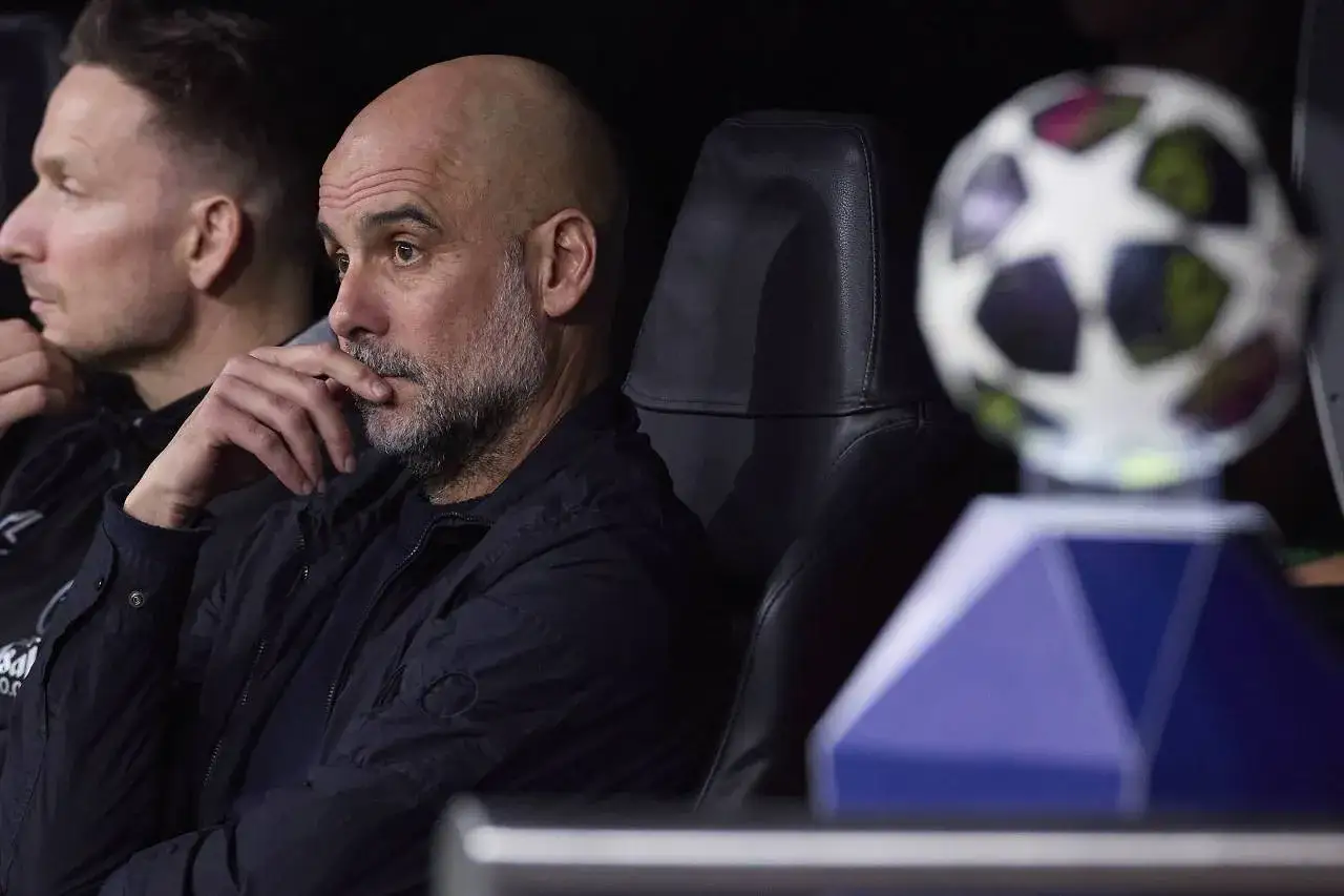 Pep Guardiola, con su caracter&iacute;stica calva y barba gris, reflexiona junto a un bal&oacute;n de la Champions League. Su edad se refleja en su serena mirada.