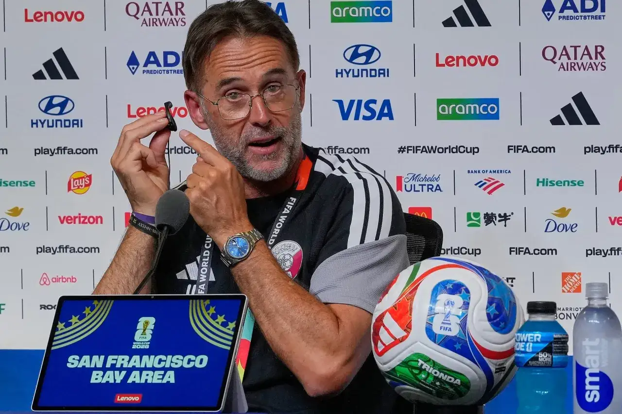 El entrenador de Qatar, con gafas, habla mientras sostiene un auricular. A su lado, un balón de fútbol y una botella de agua.