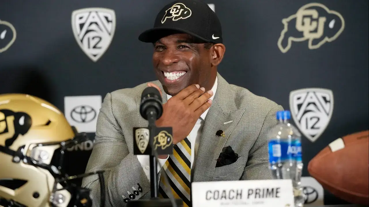 Entrenador Prime sonríe en conferencia de prensa, con un casco de fútbol americano y el logo de Pac-12 de fondo.