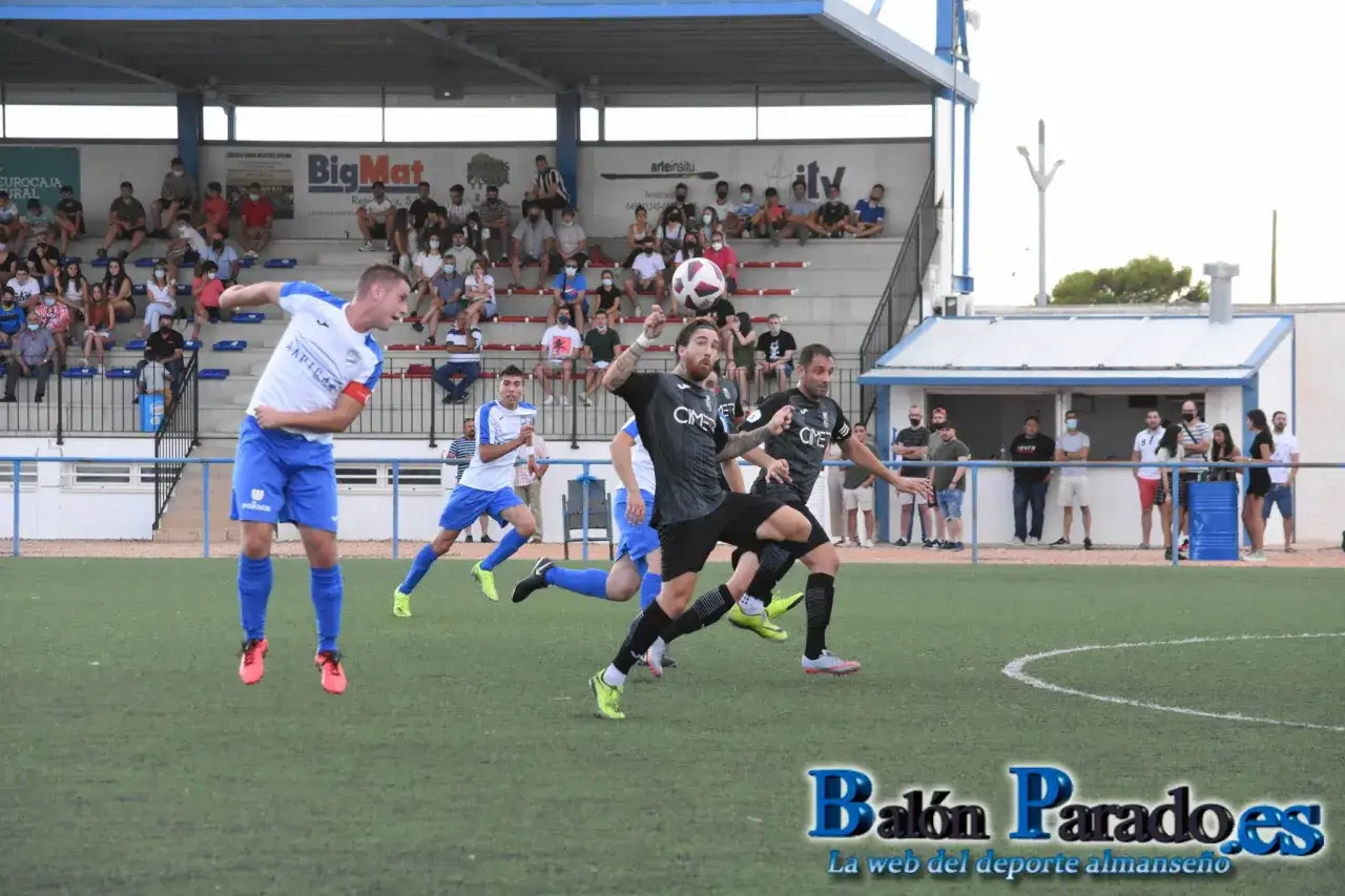 Jugadores de fútbol en acción, uno salta para cabecear el balón. El partido se desarrolla ante espectadores en las gradas. munera cf.