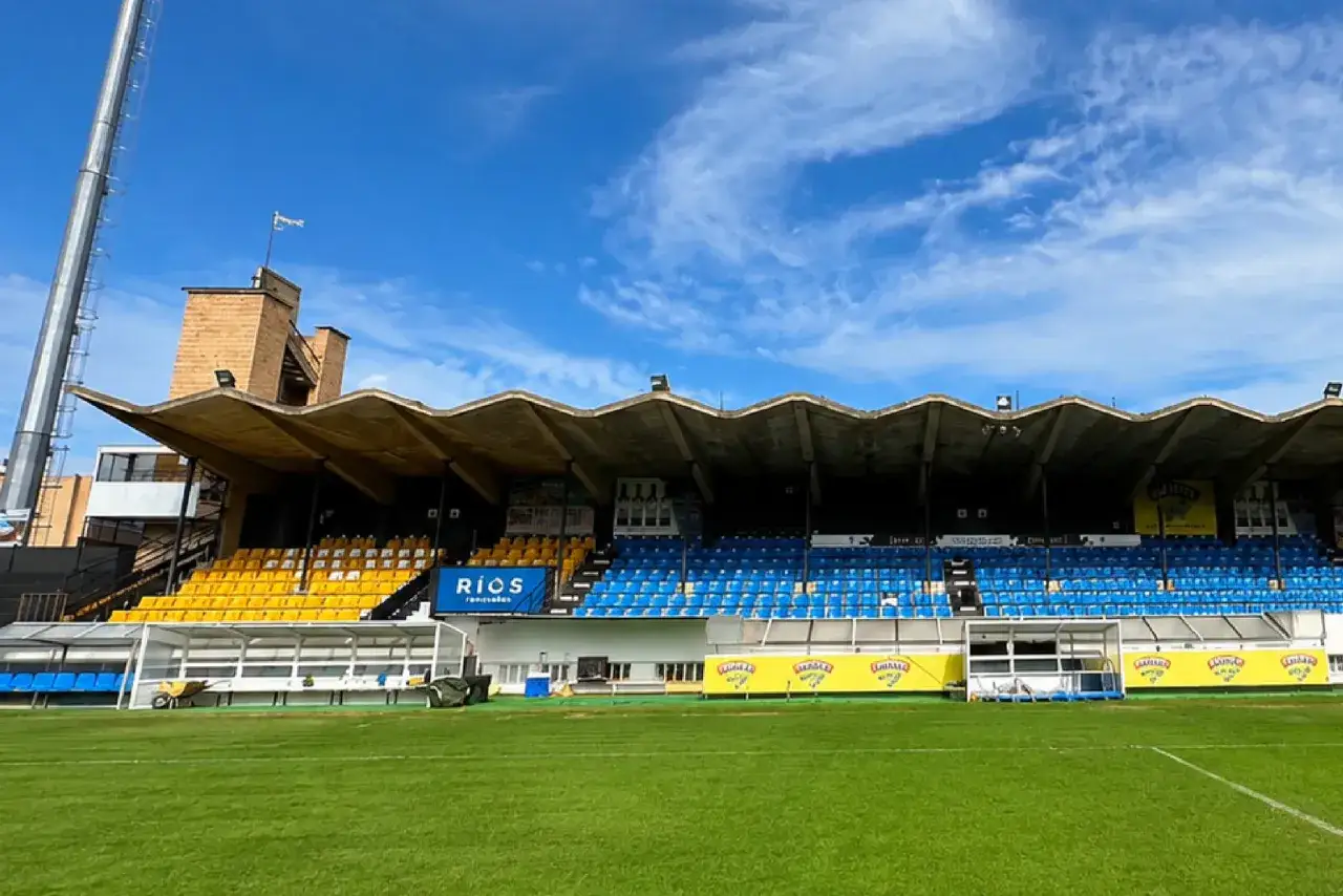 Grada de estadio con asientos azules y amarillos, lista para un partido pretemporada. Césped verde impecable.