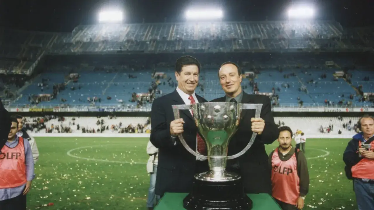 Rafa Benítez, sonriendo, sostiene un trofeo junto a otro hombre en un estadio. El fondo muestra gradas llenas y confeti en el campo.