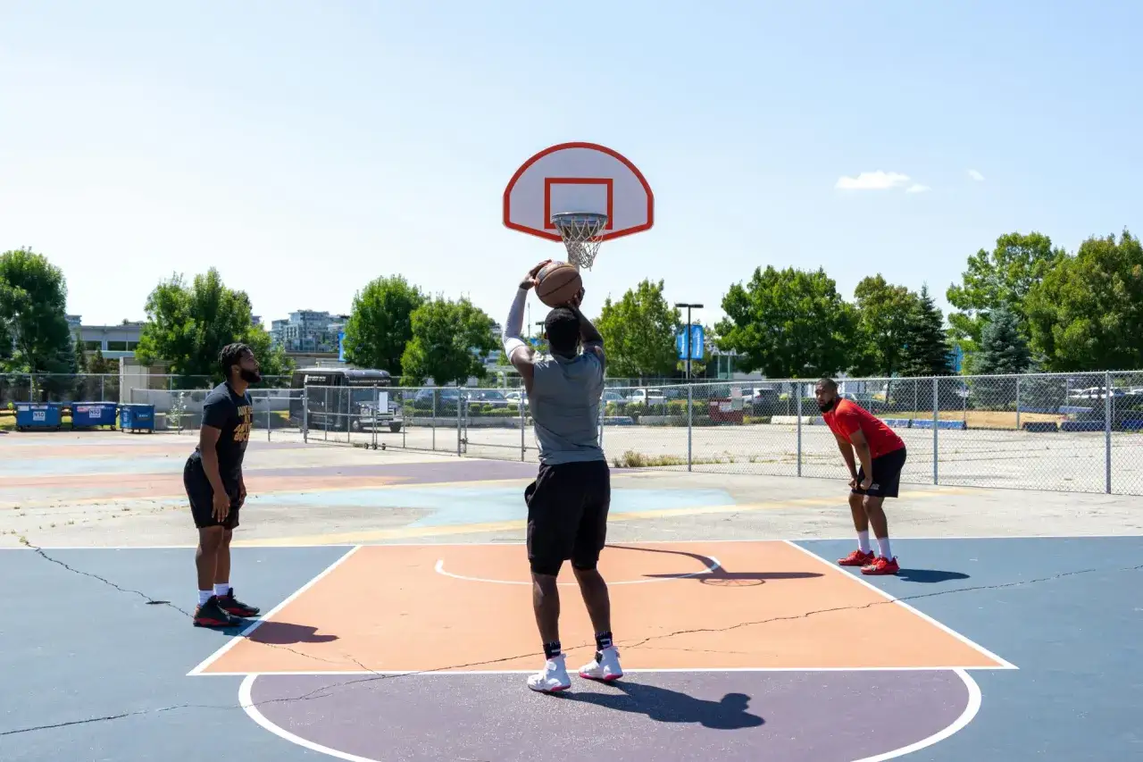 Tres hombres juegan baloncesto en una cancha exterior. Uno lanza el bal&oacute;n, mientras los otros esperan. Practican sus tiros libres.