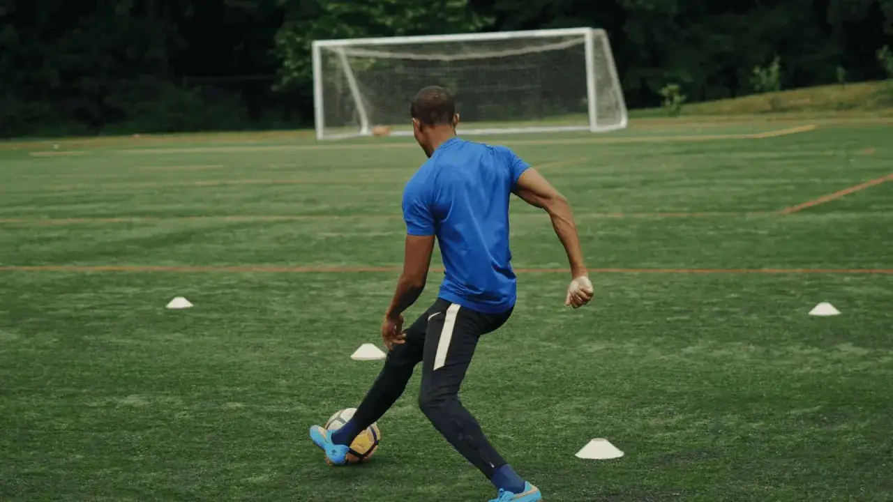 Un futbolista practica sus football skills en un campo de césped artificial, driblando conos hacia la portería.