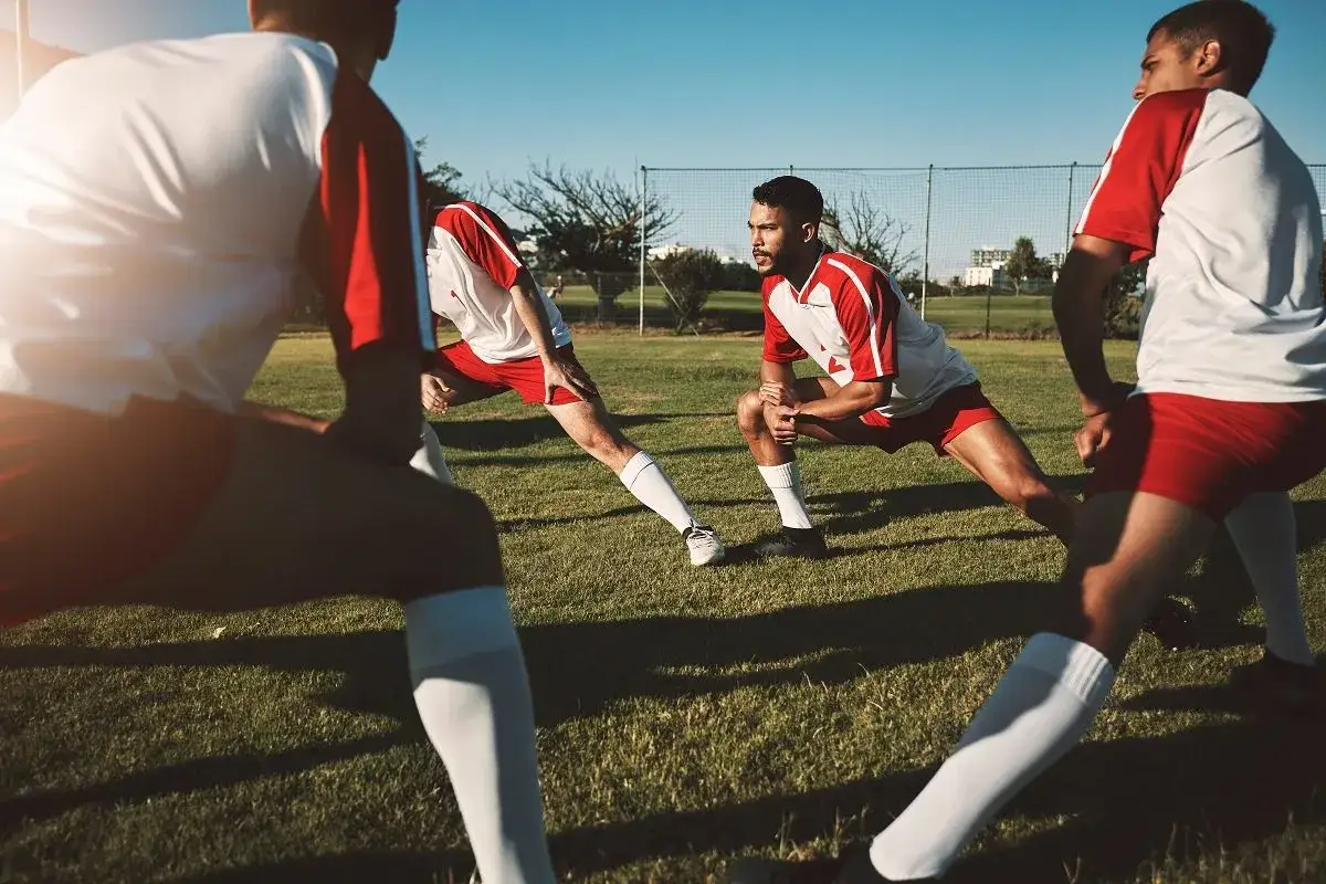 Jugadores de f&uacute;tbol realizan 20 ejercicios de calentamiento en el campo, estirando sus piernas bajo el sol.