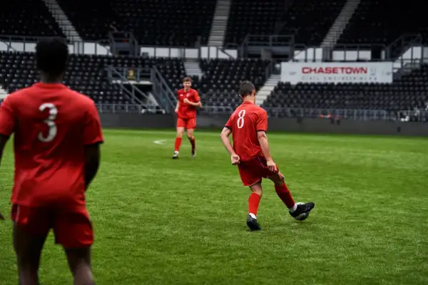 Jugadores de fútbol en uniforme rojo practicando ejercicios de pases en un campo verde, con gradas vacías al fondo.