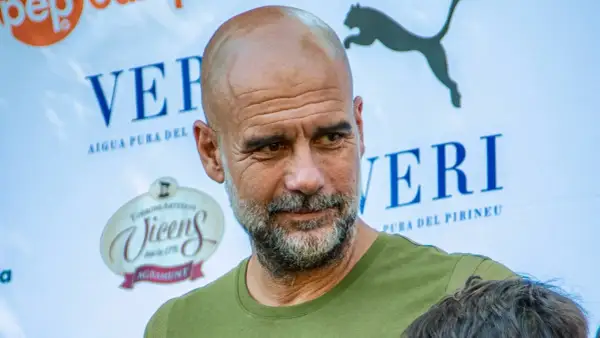 Pep Guardiola, con su mirada reflexiva, parece estar en un evento de "pep guardiola estudios". Detrás, logos de "Veri" y "Vicens".