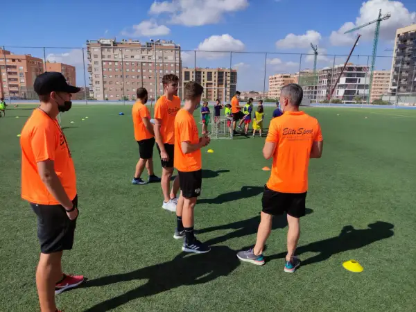 Entrenadores con camisetas naranjas dirigen a jóvenes deportistas en un campo de fútbol. Uno de ellos, con gorra negra, parece estar explicando algo, quizás sobre la equivalencia de técnico deportivo nivel 1.