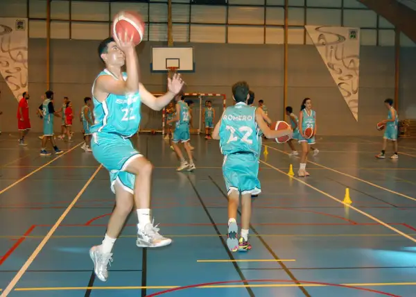 Jugadores de baloncesto realizan ejercicios analíticos en cancha. Uno salta para lanzar, otro dribla.