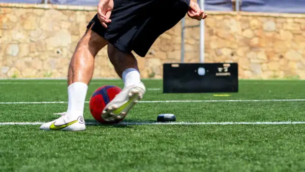 Jugador de fútbol entrenando ejercicios de velocidad en campo artificial, con balón y dispositivo de medición.