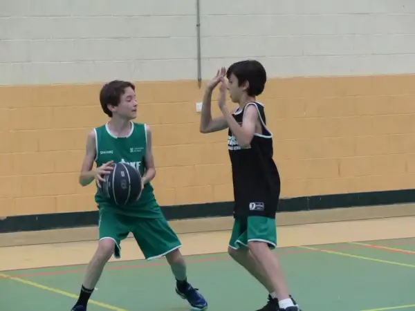 Dos niños juegan al baloncesto. Uno, con camiseta verde, se prepara para hacer un pase de pecho. El otro, con camiseta negra, se defiende.