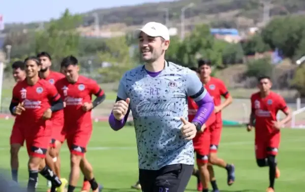 Entrenador sonriente lidera a jugadores en un partido pretemporada. Corren en un campo verde bajo el sol.