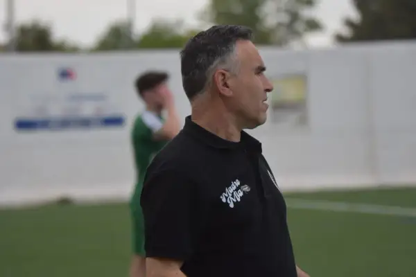 Entrenador de fútbol con polo negro, mirando hacia la derecha, con un jugador de verde al fondo.