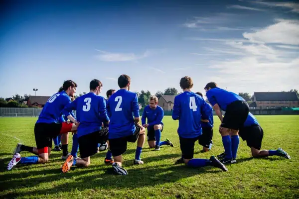 Equipo de fútbol con camisetas azules, arrodillados en el campo, preparándose para juegos de activación.