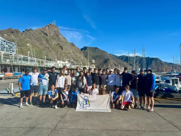 Grupo de jóvenes y adultos posan con una bandera del **CD Urda** en un puerto deportivo, con montañas y barcos de fondo.