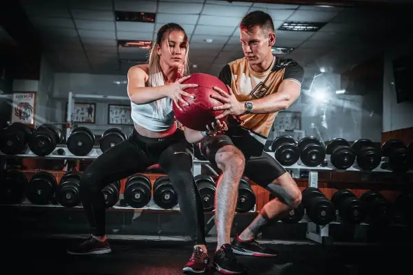 Pareja realizando ejercicios en pareja con pelota en un gimnasio, rodeados de pesas.