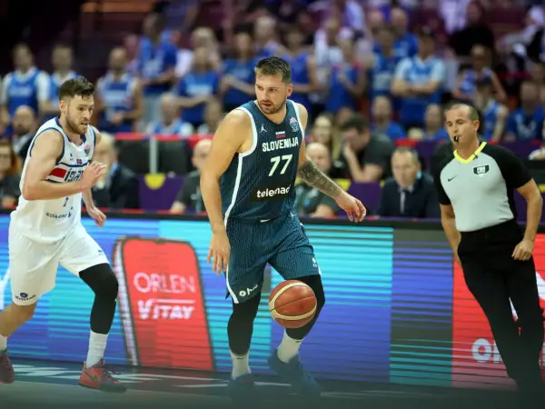 Jugador esloveno con el número 77 dribla el balón, mientras un árbitro observa atentamente. El jugador islandés, con el número 7, se prepara para defender. Se percibe una falta en ataque en el baloncesto.