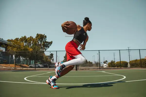 Jugadora de baloncesto en movimiento, con el balón en las manos, lista para anotar. Sus aleros de baloncesto brillan bajo el sol.