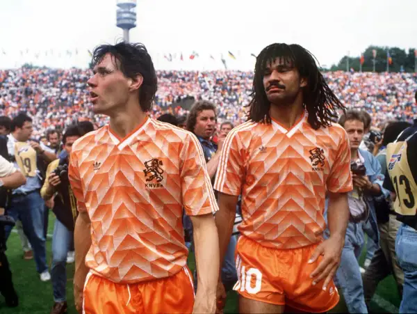 Dos futbolistas de equipos holandeses con camisetas naranjas icónicas, rodeados de aficionados y cámaras.