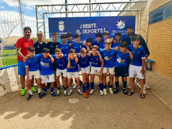 Equipo de fútbol infantil posa sonriente. El cartel de fondo anuncia "¡Siente el deporte!" y el logo del Ayuntamiento de San José.